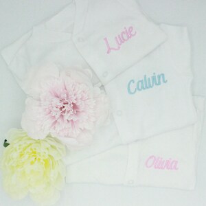 Puede incluir: Bodys blancos para bebés con nombres bordados: "Lucie" en rosa, "Calvin" en azul claro y "Olivia" en rosa. Los bodys están dispuestos con dos grandes flores en tonos rosa y amarillo.