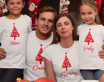 May include: Four people wearing white t-shirts with a red Christmas tree design. The shirts have names printed below the tree: Adam, Daddy, Mummy, and Emily. The family is posing in front of a fireplace decorated for Christmas.
