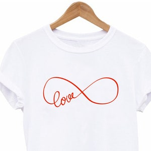 May include: White t-shirt featuring a red infinity symbol with the word "love" written in cursive. The t-shirt is displayed on a wooden hanger.