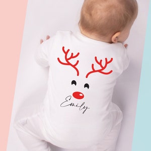 May include: White baby onesie with a red reindeer face design on the back. The design includes red antlers, black eyes, and a red nose. The name "Emily" is written in a cursive font below the face. The onesie is for a baby.