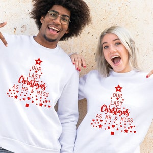 May include: Two white sweatshirts with the text "OUR LAST Christmas AS MR & MISS" in red, designed as a Christmas tree. The sweatshirts are worn by a man and a woman, both smiling and pointing at the design.