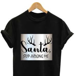 Peut inclure: T-shirt noir avec l'inscription "Santa, STOP JUDGING ME" dans un rectangle argenté, avec des graphiques de bois de cerf au-dessus. Le t-shirt est présenté sur un cintre en bois sur fond blanc.