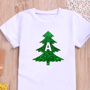 May include: White t-shirt featuring a green glitter Christmas tree design with the letter "A" in white. The shirt has short sleeves and a crew neck. The design is centered on the front of the shirt.