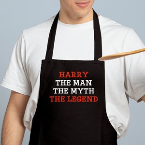 May include: Black apron with the text "HARRY THE MAN THE MYTH THE LEGEND" in white and red lettering. The apron is worn by a person wearing a white t-shirt.