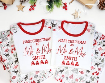 May include: Matching white and red Christmas pajamas sets. The tops have red trim and the text "FIRST CHRISTMAS as Mr & Mrs SMITH" in red. The pants have a pattern of deer, trees, and snowflakes. Festive holiday apparel.