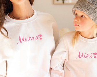 May include: Matching white sweatshirts with pink text that reads "Mama" and "Mini" with heart accents. The sweatshirts are a matching set, perfect for a mother and child. The image shows a woman and a young girl wearing the sweatshirts.