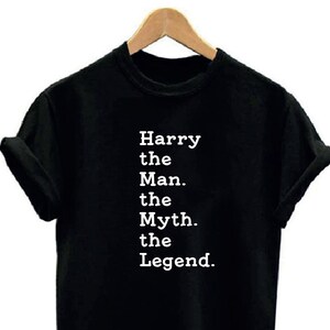 May include: Black t-shirt with the text "Harry the Man. the Myth. the Legend." in white, displayed vertically. The shirt is hanging on a wooden hanger against a white background. The sleeves are cuffed.