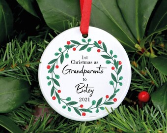 Peut inclure: Une boule de Noël blanche avec un ruban rouge. L'ornement présente une couronne verte et rouge entourant le texte "1st Christmas as Grandparents to Betsy 2021". L'ornement est entouré de feuillage vert.