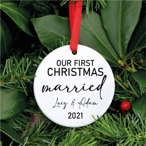 Peut inclure: Une boule de Noël blanche et ronde avec un ruban rouge. Elle porte l'inscription "OUR FIRST CHRISTMAS married Lucy & Adam 2021" en noir. L'ornement est suspendu parmi le feuillage vert.