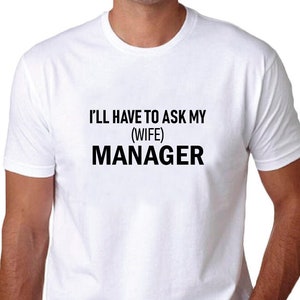 Peut inclure: T-shirt blanc avec l'inscription en noir "I'LL HAVE TO ASK MY (WIFE) MANAGER". Le t-shirt est à manches courtes et semble être fait d'une matière douce. La personne porte un jean bleu.