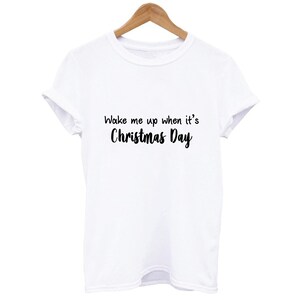 Peut inclure: T-shirt blanc avec le texte "Wake me up when it's Christmas Day" en script noir. Le t-shirt est suspendu à un cintre en bois. Le t-shirt a des manches retroussées.