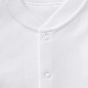May include: Close-up of a white garment with a rounded neckline and snap closures. The fabric appears to be a soft, knit material. The garment is plain white, with no visible patterns or designs.