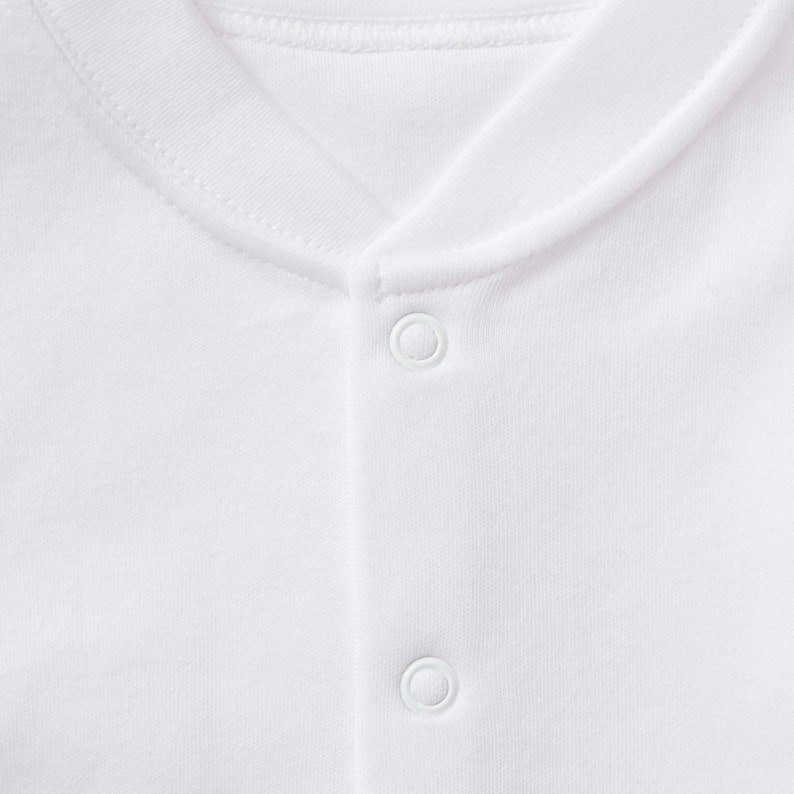 May include: Close-up of a white garment with a rounded neckline and snap closures. The fabric appears to be a soft, knit material. The garment is plain white, with no visible patterns or designs.