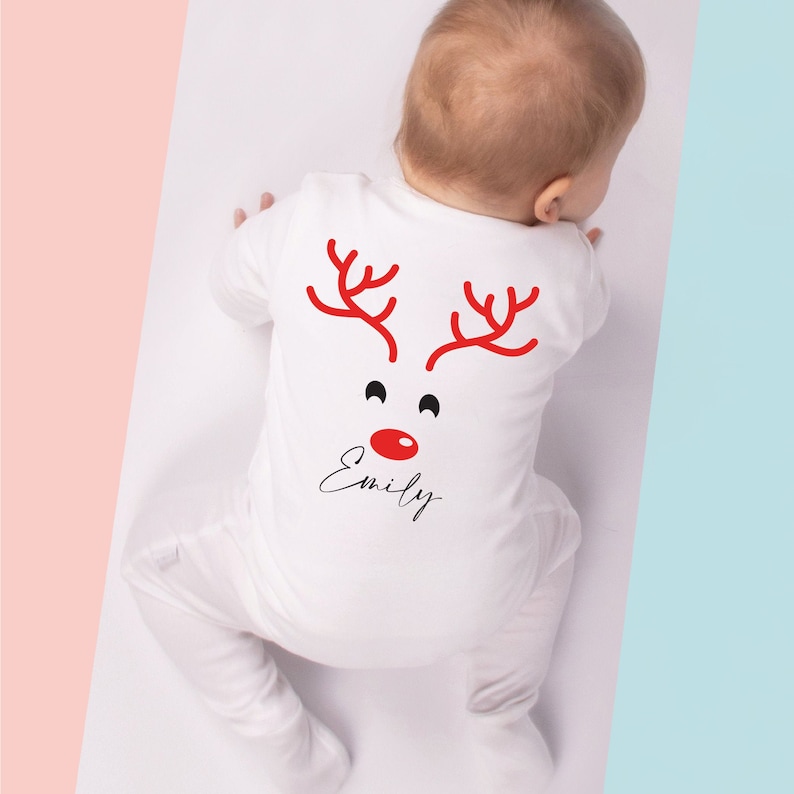 May include: White baby onesie with a red reindeer face design on the back. The design includes red antlers, black eyes, and a red nose. The name "Emily" is written in a cursive font below the face. The onesie is for a baby.