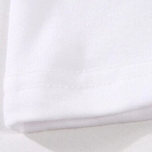 May include: Close-up of a white fabric, likely a t-shirt or similar garment. The image highlights the texture of the material and the stitching along the hem, showcasing the garment's construction and design. The fabric appears to be a soft, knit material.