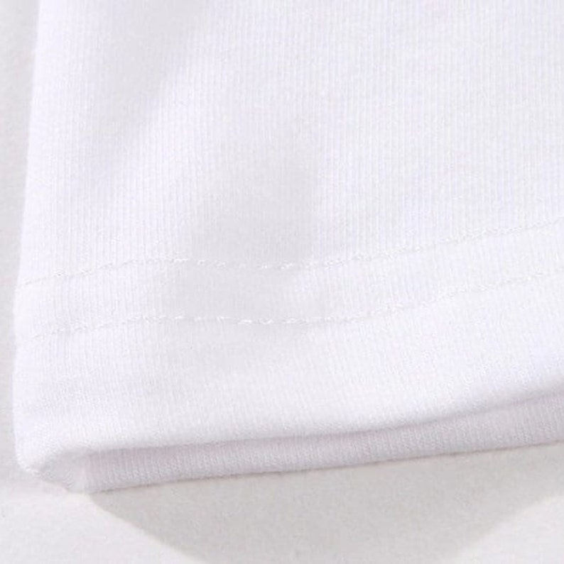 May include: Close-up of a white fabric, likely a t-shirt or similar garment. The image highlights the texture of the material and the stitching along the hem, showcasing the garment's construction and design. The fabric appears to be a soft, knit material.