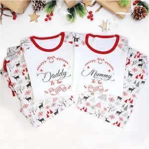 May include: Matching white Christmas pajamas with red trim and festive patterns. The shirts read "Daddy to be 2025" and "Mummy to be 2025" in a decorative font. The pajama pants feature a winter forest design with deer and snowflakes.