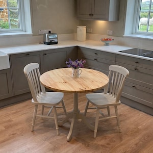 May include: A kitchen featuring light grey cabinets, white worktops, and a round wooden table with two white chairs. A vase of purple flowers sits on the table. The room has two windows and a farmhouse sink.