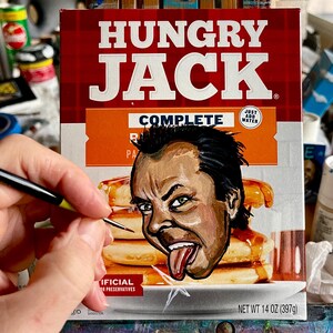May include: A Hungry Jack pancake mix box with a painted portrait of a man sticking out his tongue. The box is red and white with the words "HUNGRY JACK" in large white letters. The net weight is 14 oz (397g).