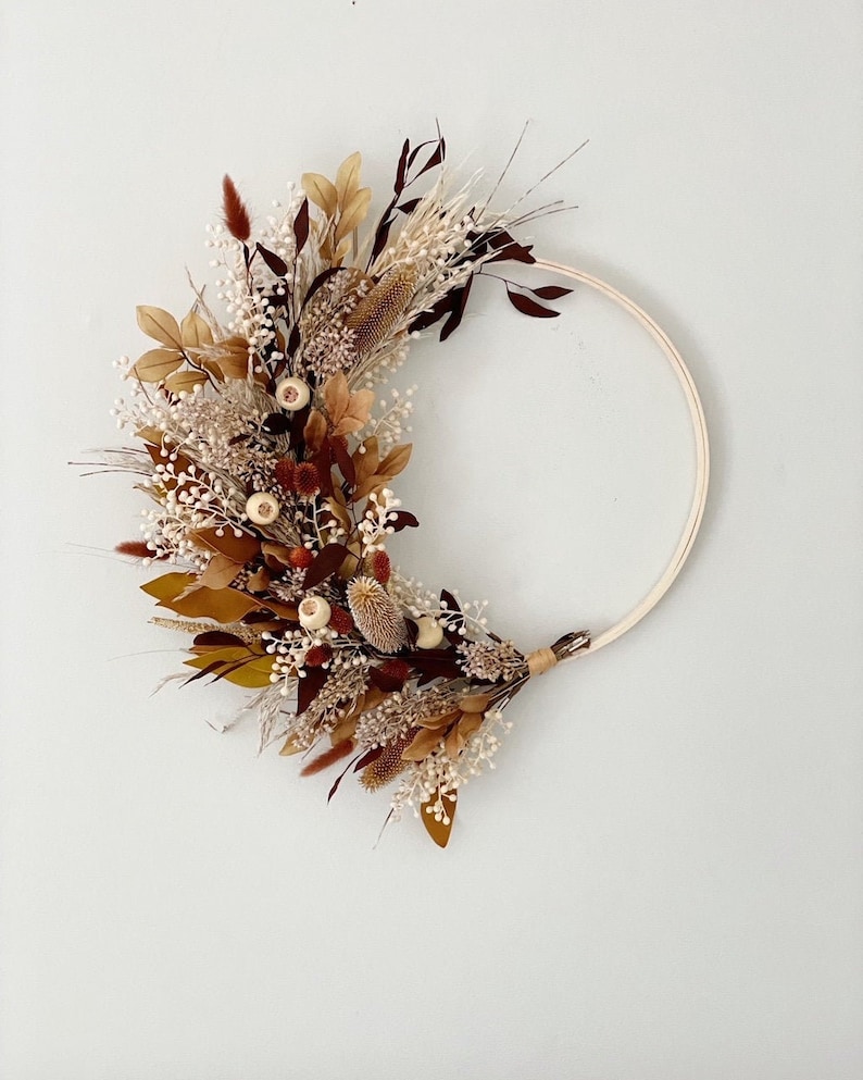 May include: A dried floral wreath with a wooden hoop. The wreath is made of dried flowers in shades of brown, white, and red. The wreath is a half circle and is hanging on a white wall.