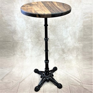 Pu&ograve; includere: Un tavolo da bistrot in metallo nero con un piano rotondo in legno. Il tavolo ha una base a tre gambe con dettagli intricati.