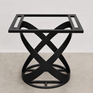 May include: A matte black metal table base featuring a square top frame with four mounting points. The central support structure consists of two curved, intersecting bands that form an X-shape, connecting the square top to a circular base ring.