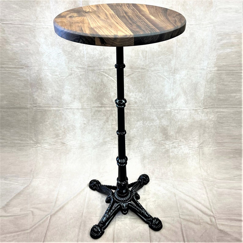 Pu&ograve; includere: Un tavolo da bistrot in metallo nero con un piano rotondo in legno. Il tavolo ha una base a tre gambe con dettagli intricati.