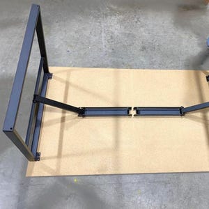 May include: Black metal table legs with a folding mechanism. The legs are attached to a light brown tabletop.