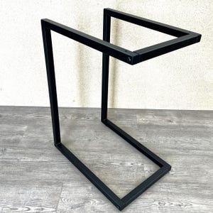 May include: A black metal side table with a geometric, open-frame design. The table features a rectangular base and a top surface, connected by a vertical support. The minimalist design is suitable for various interior styles.