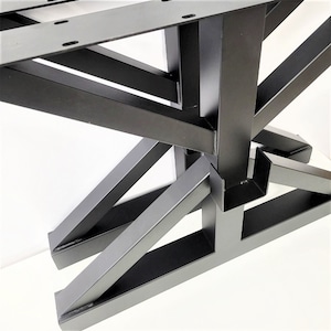 May include: Close-up of a black metal table base. The base features a geometric design with angled supports and a sturdy, industrial aesthetic. The metal is painted black, and the design suggests a modern or minimalist style.