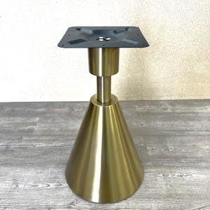 May include: A gold-colored metal table base with a square top and a conical base. The base has a cylindrical support and a black square plate with screw holes. The base is designed for a table.