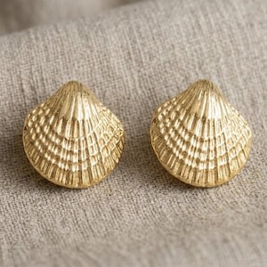 Könnte beinhalten: Zwei goldfarbene Ohrstecker in Form von Muscheln. Die Ohrringe haben eine detaillierte, strukturierte Oberfläche mit einem geriffelten Muster. Sie werden auf einem neutral getönten, strukturierten Stoffhintergrund präsentiert.