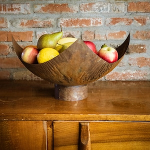 Może przedstawiać: Rustykalna metalowa miska z szerokim, rozszerzającym się brzegiem stoi na drewnianej powierzchni. Miska jest wypełniona różnymi owocami, w tym jabłkami, gruszkami i cytrynami.