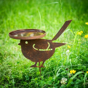 Puede incluir: Un comedero para pájaros de metal oxidado con un plato poco profundo para agua o semillas. El comedero está diseñado para parecer un pájaro con una cola larga y una cabeza redonda. El comedero está de pie en un área cubierta de hierba.