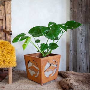 Op de afbeelding: Een grote groene Monstera-plant in een vierkante, roestige metalen pot met een uitgesneden ontwerp van bomen. De pot heeft een rustieke, verweerde uitstraling.