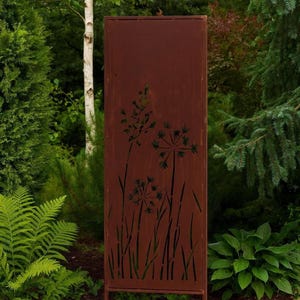 A rust-colored metal fence with a design of flowers stands in a lush, green garden surrounded by various plants and trees.