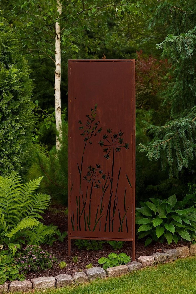 A rust-colored metal fence with a design of flowers stands in a lush, green garden surrounded by various plants and trees.