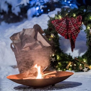 May include: A rusty metal fire pit with a small fire burning inside. The fire pit is sitting on a snowy surface. There is a basket of firewood and a Christmas wreath with lights in the background.