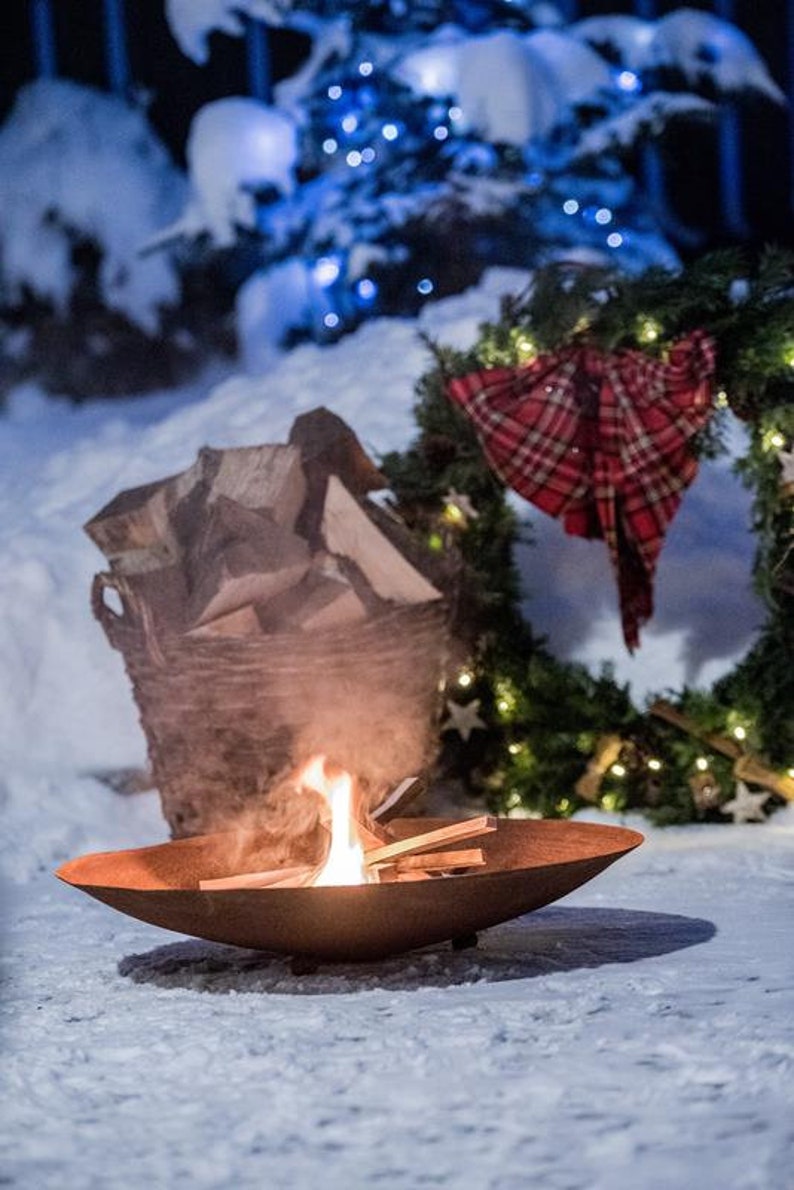 May include: A rusty metal fire pit with a small fire burning inside. The fire pit is sitting on a snowy surface. There is a basket of firewood and a Christmas wreath with lights in the background.