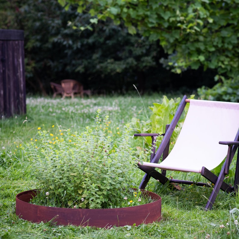 A garden with a white lawn chair and a round planter filled with green plants and yellow flowers, surrounded by a wooden fence and trees.