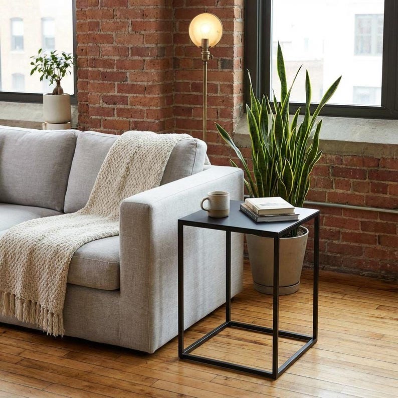 a cozy living room with a gray sofa, a black metal side table, and a potted plant. The sofa is draped with a white blanket, and there is a coffee mug and a book on the side table. The room has a brick wall and a window, creating a warm and inviting atmosphere.