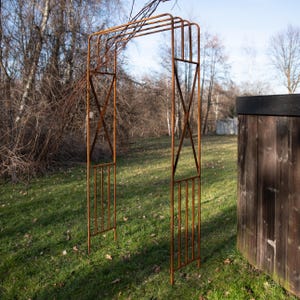 An orange metal arch stands in a grassy area, with a wooden fence and bare trees in the background.