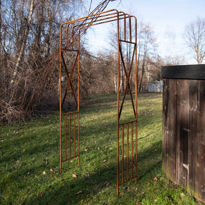 An orange metal arch stands in a grassy area, with a wooden fence and bare trees in the background.