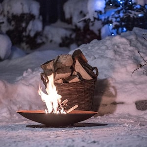 Può includere: Un focolare con un piccolo fuoco che brucia all'interno, circondato da neve. Un cesto di vimini pieno di legna da ardere si trova accanto al focolare.