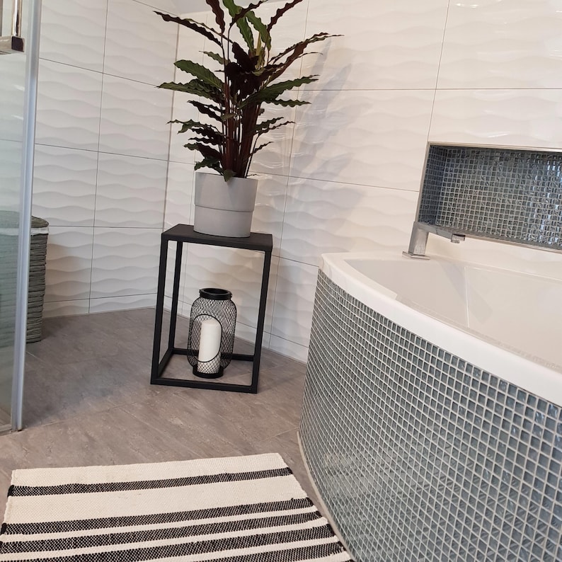 a bathroom with a white bathtub, a black and white striped rug, a potted plant on a black stand, and a gray and white striped rug.