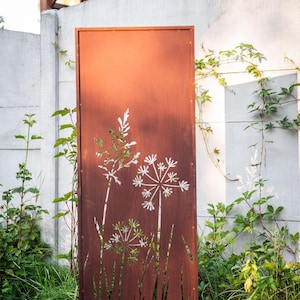 a rust-colored metal gate with a white floral design on it, standing in a grassy area with a white wall and a wooden trellis structure in the background.