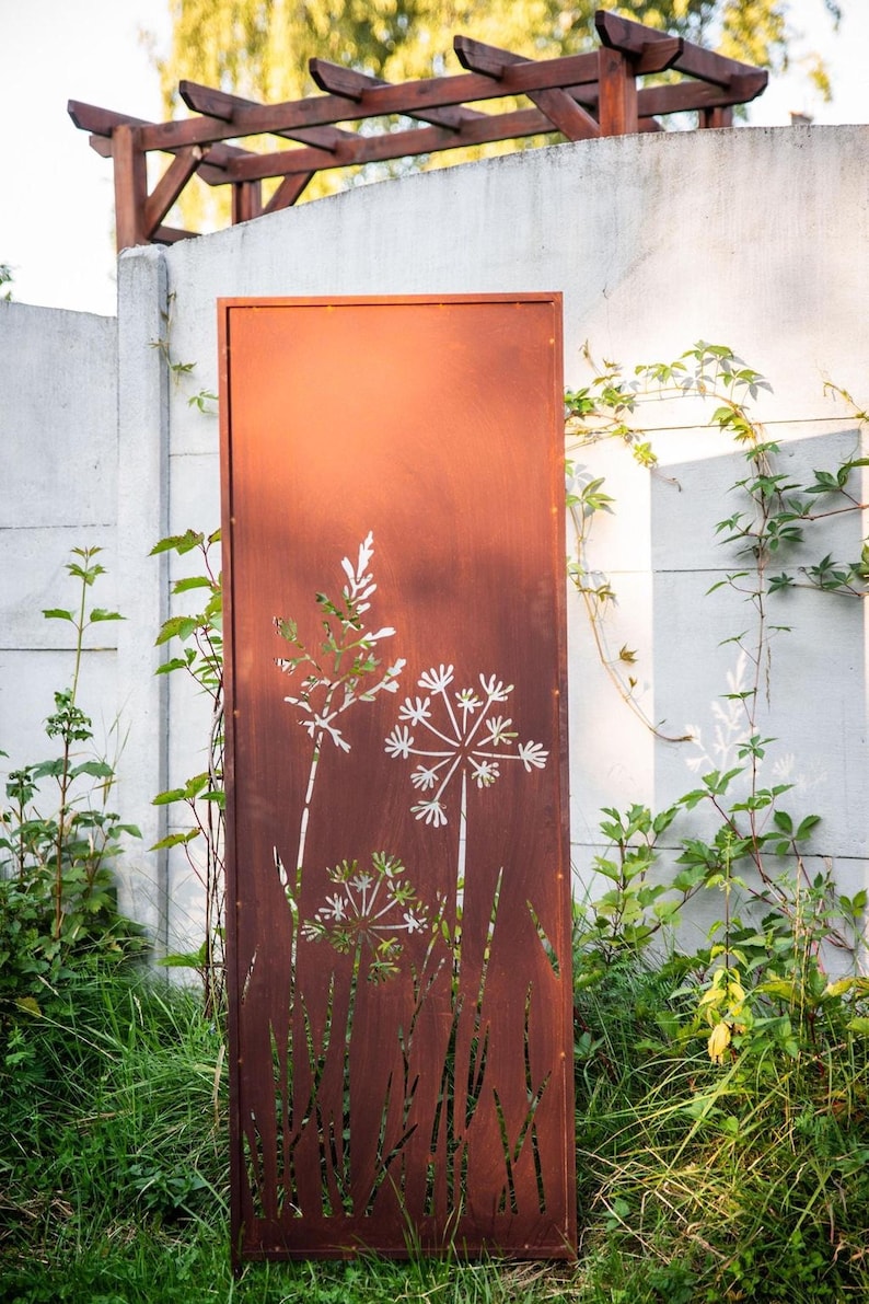 a rust-colored metal gate with a white floral design on it, standing in a grassy area with a white wall and a wooden trellis structure in the background.