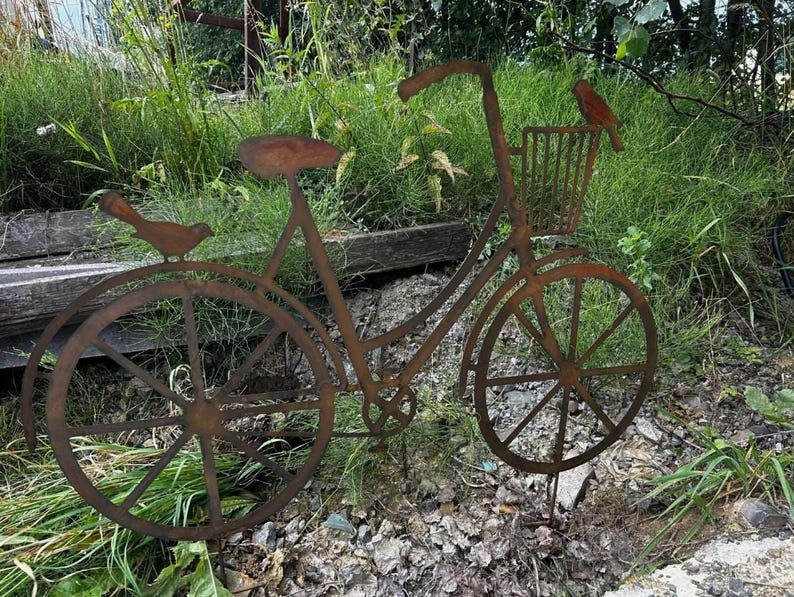 A rusted metal bicycle sculpture stands in a grassy area, with a bird perched on the handlebars.