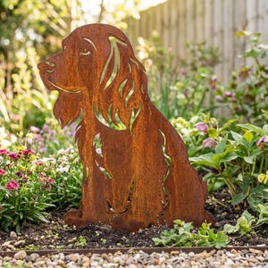 Könnte beinhalten: Rostbraune Metall-Gartenskulptur in Form eines sitzenden Hundes. Das Kunstwerk weist Aussparungen im Fell auf. Die Dekoration steht in einem Beet mit Blumen und Grünpflanzen und dient als dekoratives Element im Freien.