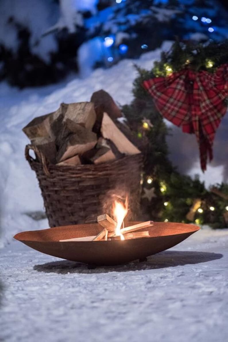 May include: A rusty metal fire pit with a small fire burning inside. The fire pit is sitting on a snowy surface. A wicker basket filled with firewood is in the background.